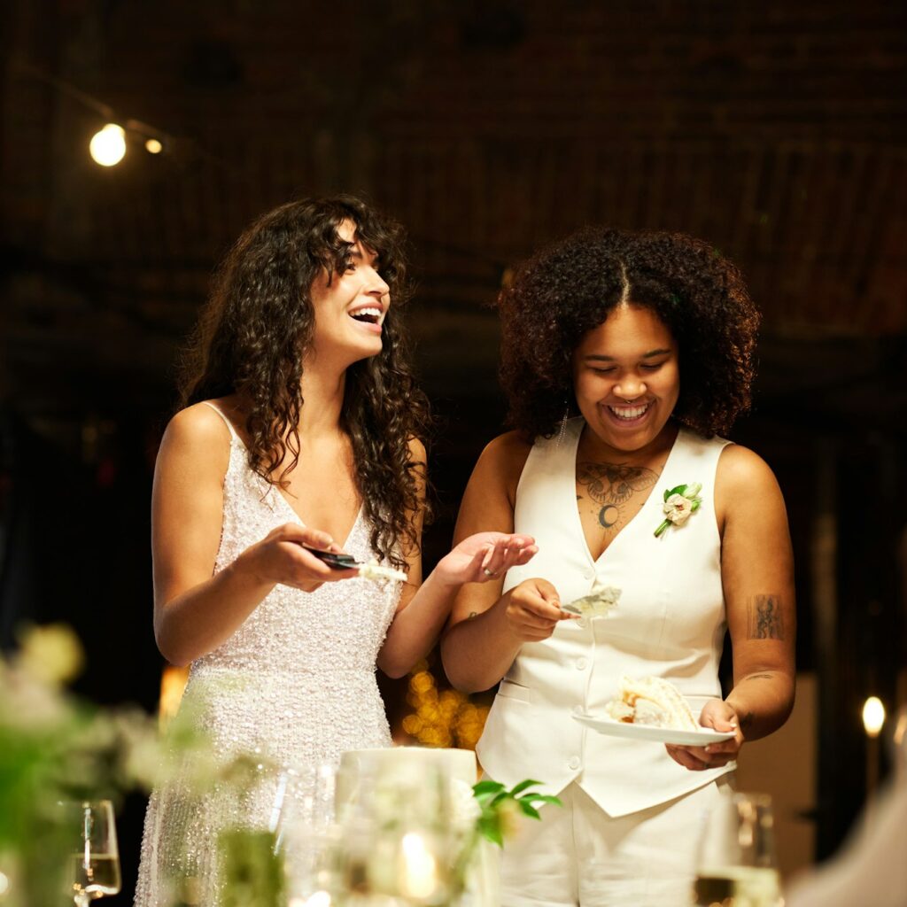 Cheerful young lesbian brides in wedding attire spreading cake among guests